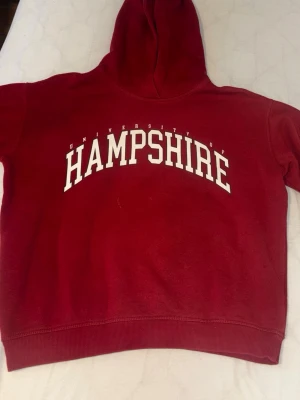 Röd Hampshire hoodie med tryck - Snygg röd hoodie med vit text 'University of Hampshire' tryckt över bröstet. Klassisk modell med huva och ribbade muddar vid ärmslut och nederkant. Perfekt för dig som gillar college-stil. Mjuk och skön bomullsblandning.