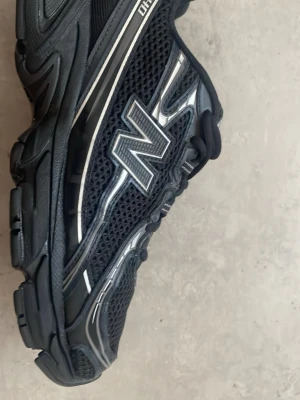 Svarta New Balance 2002R sneakers - Snygga svarta New Balance 2002R sneakers med mesh och syntetdetaljer. Skorna har reflekterande silverdetaljer och den klassiska N-loggan på sidan. Tjock sula för extra komfort och sportig look. Perfekta för dig som gillar streetwear och stilrena sneakers.