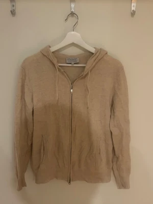 Beige hoodie med dragkedja - Beige hoodie från Ahlquist Clothing i storlek S. Mjuk och skön med dragkedja framtill, två fickor och ribbade muddar. Klassisk huva med dragsko. Perfekt för en chill och avslappnad stil.