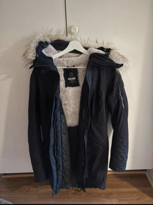 Svart parkasjacka från 8848 Altitude - Snygg svart parkasjacka från 8848 Altitude med avtagbar huva och fuskpäls. Jackan har dragkedja, flera fickor och mysigt vitt teddyfoder på insidan. Perfekt för kalla dagar och har en modern, längre passform.