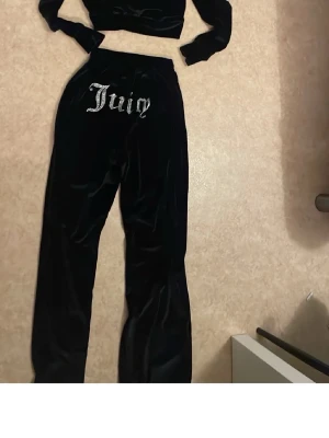 Juicy Couture svart velourset - Juicy Couture loungewear-set i svart velour med glittrande 'Juicy'-text på både byxorna och hoodien. Setet består av en croppad hoodie med långa ärmar och ett par mjuka byxor med resår i midjan. Perfekt för en chill och trendig look.