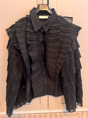 Svart skjorta med volangdetaljer - Unik svart skjorta från Copenhagen Muse med flera lager av volangliknande plisserade detaljer över hela plagget. Skjortan har lång ärm och är tillverkad i ett tunt, transparent tyg som ger en cool och edgy look.