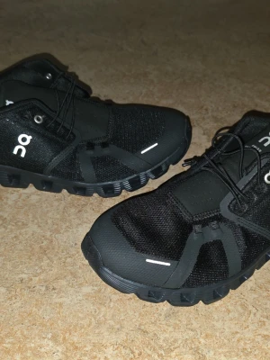 Svarta On Running sneakers - Snygga svarta sneakers från On Running med mesh- och syntetmaterial, vita loggor på sidan och tungan samt unika snörningar. Skorna har en sportig design och grov sula som ger bra grepp. Perfekta för dig som gillar stilrena och funktionella sneakers.