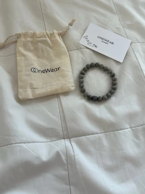 Grått pärlarmband från OneWear - Snyggt armband från OneWear med runda grå pärlor i stenlook och en svart detaljpärla. Armbandet är elastiskt och levereras med en tygpåse med OneWear-logga. Perfekt accessoar för att lyfta din stil.