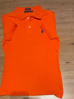 Orange pikétröja från Ralph Lauren - Snygg och klassisk orange pikétröja från Ralph Lauren med korta ärmar och blå logga broderad på bröstet. Tröjan har krage, två knappar framtill och är gjord i mjuk bomull. Perfekt för en avslappnad men ändå stilren look. Står att den är storlek s men är mindre!