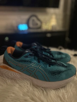 Blå ASICS sneakers med orange detalj - Säljer ett par snygga blå ASICS sneakers med vita och svarta detaljer. Skorna har en låg profil, mesh-överdel för bra ventilation och vit sula med PureGEL-teknologi för extra komfort. Perfekta för dig som gillar sportig stil eller använda för träning. Ny pris på dessa ligger ca/1400kr   Jag säljer dessa eftersom jag inte har användning för dom längre, väldigt sköna skor att springa/träna hör av er vid andra tankar 