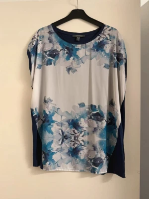 Blommig topp i blå och vitt - Snygg topp med blommigt mönster i blå och vita toner. Toppen har rund halsringning, ärmlös design och löst sittande passform. Materialet känns mjukt och lätt, perfekt för varmare dagar. Blå detaljer längs sidorna ger en cool kontrast.