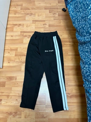 Palm Angels byxor - Säljer ett par svarta mjukisbyxor från Palm Angels med vita ränder längs sidorna och logga tryckt framtill. Byxorna har avslappnad passform och resår i midjan för extra komfort. Perfekta för en chill och trendig look. Felfria inga skador eller liknande.