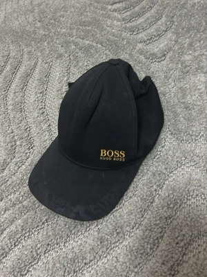 Svart keps från Hugo Boss - Snygg svart keps från Hugo Boss med guldigt BOSS-tryck framtill. Klassisk böjd skärm och justerbar baksida. Materialet är troligtvis bomull eller polyester, perfekt för en stilren look.