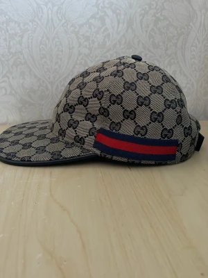 Gucci keps med GG-mönster och röd-blå rand - Snygg keps från Gucci i beige och svart med klassiskt GG-monogram över hela kepsen. På sidan finns en ikonisk röd och blå rand. Kepsen har böjd skärm och justerbar rem bak. Materialet är canvas med detaljer i textil.