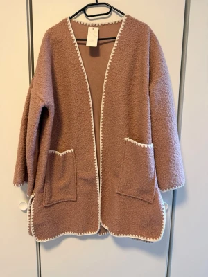 Rosa teddycardigan med vita kanter - Mysig rosa teddycardigan med två stora fickor framtill och dekorativa vita kanter längs öppning, ärmslut och nederkant. Jackan har en avslappnad passform och är perfekt för lager-på-lager. Materialet är fluffigt och ger en trendig look.