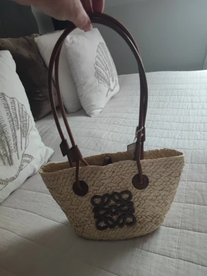 Stråväska med bruna läderhandtag - Snygg shoppingväska i flätad strådesign från Loewe med bruna läderhandtag och dekorativt brunt lädermärke framtill. Perfekt för dig som gillar en naturlig och trendig look. Rymlig modell som rymmer det mesta du behöver under dagen.