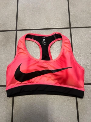 Rosa sport-bh från Nike - Snygg sport-bh från Nike i stark rosa färg med svart resår och stort svart Nike-logga framtill. Racerback-modell för bra stöd och rörelsefrihet. Tillverkad i ett stretchigt och snabbtorkande material som passar perfekt till träning.