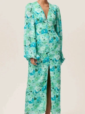 Adoore Grön blommig långklänning med slits - Adoore Snygg långklänning med grönt och blått blommönster, djup v-ringning och långa puffärmar. Klänningen har knappar framtill och en hög slits som ger en trendig look. Perfekt för dig som vill sticka ut med färg och mönster.