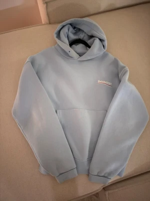 Ljusblå hoodie  - Snygg ljusblå hoodie med broderad logga på bröstet i vitt och rött. Hoodien har en klassisk känguruficka framtill, ribbade muddar och en stor huva. Perfekt för dig som gillar streetwear och vill ha något trendigt och bekvämt.