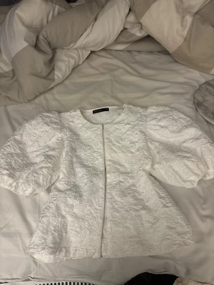 Vit puffärm blus från Zara - Supersnygg vit blus från Zara med puffiga, korta ärmar och strukturmönstrat tyg. Rak passform och rund halsringning ger en trendig look. Perfekt att styla med jeans eller kjol för en chic outfit.