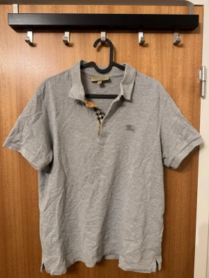 Grå pikétröja från Burberry - Klassisk grå pikétröja från Burberry med korta ärmar och den ikoniska rutiga detaljen vid knäppningen. Tröjan har en liten broderad logga på bröstet och är tillverkad i mjuk bomull. Perfekt till en avslappnad men ändå stilren look.