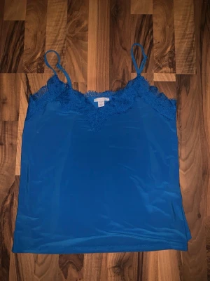 Blå linnetopp med spetsdetaljer - Snygg blå linnetopp med tunna axelband och spetsdetaljer längs v-ringningen. Perfekt att styla till jeans eller kjol för en trendig look. Materialet känns mjukt och lätt mot huden.