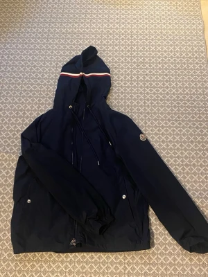 Marinblå Moncler vindjacka med huva - Snygg marinblå vindjacka från Moncler med huva och dragsko. Jackan har en vit och röd rand på huvan samt Moncler-logga på ärmen. Två fickor framtill med tryckknappar och dragkedja. Perfekt för blåsiga dagar.