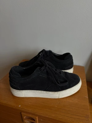Svarta sneakers från Axel Arigato - Stilrena svarta sneakers från Axel Arigato i mocka med vita, tjocka gummisulor. Klassisk låg modell med snörning och diskret logga på sidan. Liten bit av kanten som lossnat på höger häl. Annars använda ca 5 gånger och sen dess bara stått i min hall i 5 år. Storlek 37. Ingen skolåda medföljer 