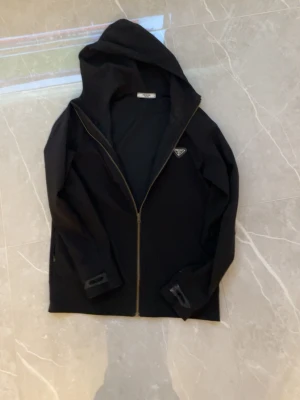 Svart hoodie med dragkedja från Prada - Snygg svart hoodie från Prada med dragkedja framtill och klassisk huva. Diskret logga på bröstet och långa ärmar. Tillverkad i ett mjukt material som känns bekvämt och stilrent. Perfekt för dig som gillar exklusiva streetwear-plagg.