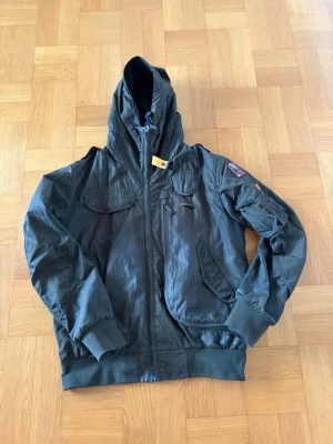 Mörkgrön vindjacka Parajumpers - Stilren mörkgrön vindjacka från Parajumpers med huva, dragkedja och flera fickor framtill. Jackan har ribbade muddar, patch på ärmen och den klassiska gula detaljen vid halsen. Perfekt för blåsiga dagar och streetwear-stil.