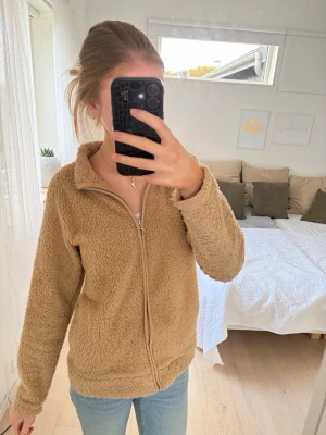 Beige teddyjacka med dragkedja - Mysig teddyjacka i beige färg med hel dragkedja framtill och hög krage. Jackan har en fluffig och mjuk textur som ger en avslappnad look. Perfekt att slänga på sig när det är kyligt ute. Passar bra till jeans och sneakers.