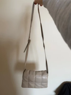 Beige axelremsväska - Säljer en beige quiltad handväska med justerbar axelrem. Väskan har en mjuk, vadderad look med rutmönster och är tillverkad i konstläder. Perfekt för dig som gillar stilrena och trendiga accessoarer.