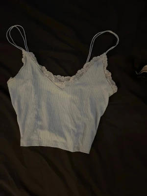 Ljusblå ribbad linnetopp med spets - Söt ljusblå linnetopp med tunna dubbel axelband och ribbad struktur. Toppen har v-ringning och dekorativ spetskant längs urringningen för en extra feminin touch. Perfekt att styla med jeans eller kjol.