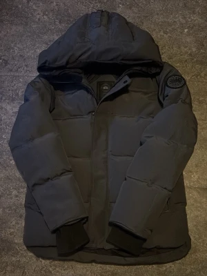 Mörk grå/svart dunjacka med huva från Canada Goose - Säljer en svart/mörk grå dunjacka från Canada Goose med stor huva och patch på ärmen. Jackan har quiltad design, ribbade muddar vid ärmsluten och dragkedja framtill. Perfekt för kalla vinterdagar och riktigt snygg streetstil.