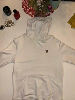 Vit hoodie från Lyle & Scott - Säljer en stilren vit hoodie från Lyle & Scott med deras klassiska gula örnlogga på bröstet. Hoodien har huva med dragsko och en stor magficka framtill. Tillverkad i mjukt bomullsmaterial som är skönt att bära.