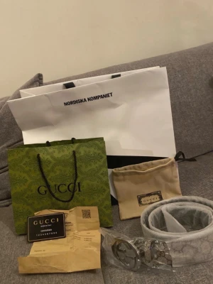 Gucci bälte med silverfärgat spänne - Säljer ett snyggt Gucci bälte med klassiskt GG-spänne i silverfärg. Bältet är ljust med diskret mönster och levereras med originalpåse, påse från Nordiska Kompaniet och kvitto. Perfekt accessoar för att lyfta din outfit.