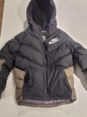 Nike dunjacka med huva svart/beige - Snygg dunjacka från Nike i svart och beige med huva och dragkedja framtill. Jackan har två fickor på sidorna och en vit Nike-logga på bröstet. Perfekt för kalla vinterdagar tack vare det tjocka fodret och den sportiga looken.