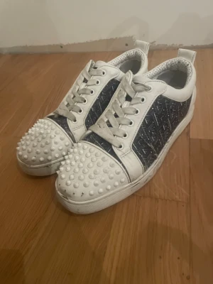 Christian Louboutin sneakers med nitar - Unika sneakers från Christian Louboutin i vitt och svart läder med mönstrade paneler och vita nitar på tån. Några nitar fattas längst fram på ena skon och några defekter. Storleken oklar pågund av att jag säljer dem åt en vän men skulle gissa på 42-43 typ.
