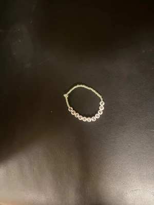 Pärlarmband med text WÅRKRÖSBARN - Sött armband med små ljusgröna pärlor och vita bokstavspärlor som stavar 'MASKROSBARN'. Perfekt accessoar för dig som gillar personliga smycken och vill ha något unikt på handleden.