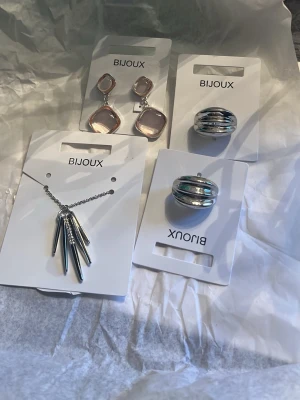 Smyckeset från Bijoux i silver och rosa - Säljer ett smyckeset från Bijoux med ett par fyrkantiga örhängen i rosa och silver, två breda silverfärgade ringar samt ett halsband med hängande stavar i silver. Perfekt för dig som gillar moderna accessoarer med stilren design. helt nya och aldrig använda. köpta i guldfynd 