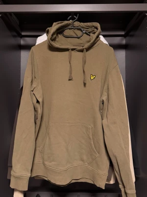 Olivgrön hoodie från Lyle & Scott - Snygg olivgrön hoodie från Lyle & Scott med klassisk gul logga på bröstet. Hoodien har huva med dragsko och en stor känguruficka framtill. Tillverkad i mjukt bomullsmaterial och har ribbade muddar vid ärmslut och nederkant. Perfekt för en avslappnad stil.