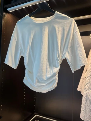 Vit croppad t-shirt  - Säljer en vit croppad t-shirt där det går in i midjan som ger en snygg rynkad effekt. T-shirten har korta ärmar och en klassisk rund halsringning. Perfekt för dig som vill ha en enkel men trendig look.