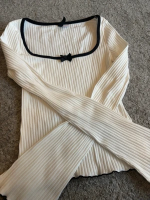 Vit ribbad topp med rosett från H&M - Supersöt vit ribbad topp från H&M med svarta detaljer och två små rosetter vid halsen. Toppen har långärmad design och en fyrkantig ringning med kontrastkant. Perfekt för att skapa en stylish look.