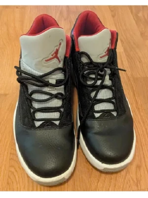 Svarta och röda Air Jordan sneakers - Snygga Air Jordan sneakers i svart och rött med vit sula. Skorna har en klassisk låg siluett, svart mesh och läder på ovandelen samt röd insida. Tungan är grå med röd Jumpman-logga. Perfekta för dig som gillar streetwear och basketstil.