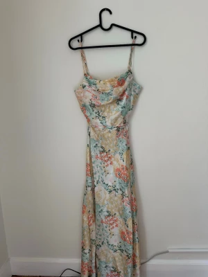 Blommig långklänning med tunna band - Säljer en långklänning med tunna axelband och vacker blommig print i pastelliga toner av beige, grön, vit och korall. Klänningen har en mjukt draperad urringning och knyts i en rosett bak i ryggen 