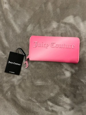 Rosa plånbok från Juicy Couture - Snygg rosa plånbok från Juicy Couture med präglad logga på framsidan och dragkedja med JC-berlock. Tillverkad i ett tåligt konstläder med struktur. Perfekt storlek för kort, cash och mynt. Kommer med originaltaggar.