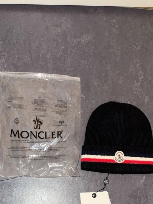Moncler mössa - Säljer en svart ribbstickad mössa från Moncler med uppvikt kant och klassisk Moncler-logga framtill. Mössan har en snygg rand i rött, vitt och blått längs kanten. Helt oanvänd.