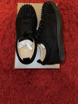 Svarta Louboutin sneakers med nitar - Snygga svarta sneakers från Christian Louboutin i mocka med svarta nitar på tån. Skorna har svarta skosnören, diskret logga på innersulan och klassisk låg siluett. Perfekta för dig som vill sticka ut med en edgy och lyxig stil.