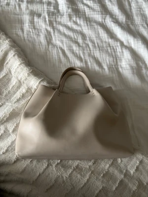 Beige väska från Zara - Snygg beige handväska från Zara med mjuk, slät yta och dubbla handtag. Väskan har en enkel och stilren design med rymligt huvudfack. Perfekt för dig som gillar en clean och modern look. Tillverkad i konstläder.