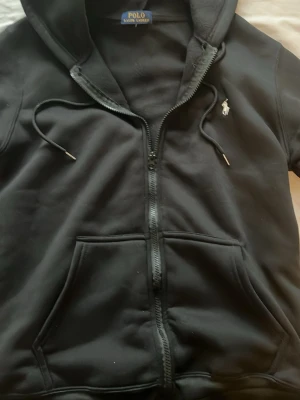 Svart hoodie från Polo Ralph Lauren - Svart hoodie med dragkedja från Polo Ralph Lauren. Klassisk design med huva och snören, två stora fickor framtill och den ikoniska vita Polo-loggan broderad på bröstet. Perfekt för en avslappnad och stilren look.