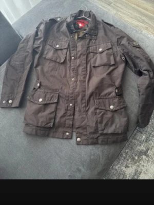 Brun vindjacka från Wellensteyn  - Snygg brun vindjacka från Fjällräven med fyra stora fickor framtill och tryckknappar. Jackan har hög krage och justerbar midja för skön passform. Tillverkad i slitstarkt material som passar perfekt för utomhusaktiviteter.