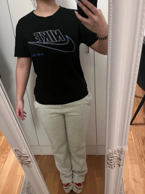 Svart Nike t-shirt med blå logga - Svart t-shirt från Nike med stor blå och vit logga och texten 'Just Do It' på bröstet. Klassisk passform med rund hals och korta ärmar. Perfekt för en sportig och avslappnad stil.