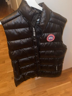 Svart dunväst från Canada Goose - Snygg svart dunväst från Canada Goose med klassisk logga på bröstet och dragkedja framtill. Västen har hög krage, två fickor med lock och quiltad design. Perfekt för lager-på-lager och håller dig varm med stil.