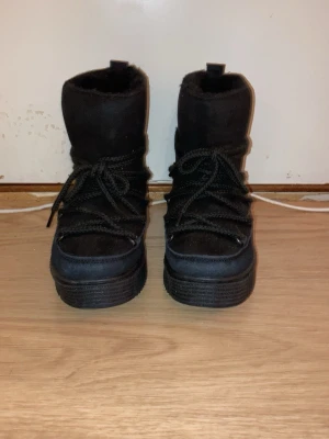 Svarta moon boots med snörning - Säljer ett par svarta moon boots med grov sula och snörning framtill. Bootsens ovandel är i ett mjukt material och de har en hög, vadderad design som håller värmen. Perfekta för kalla dagar och riktigt snygga till vinterlooken.
